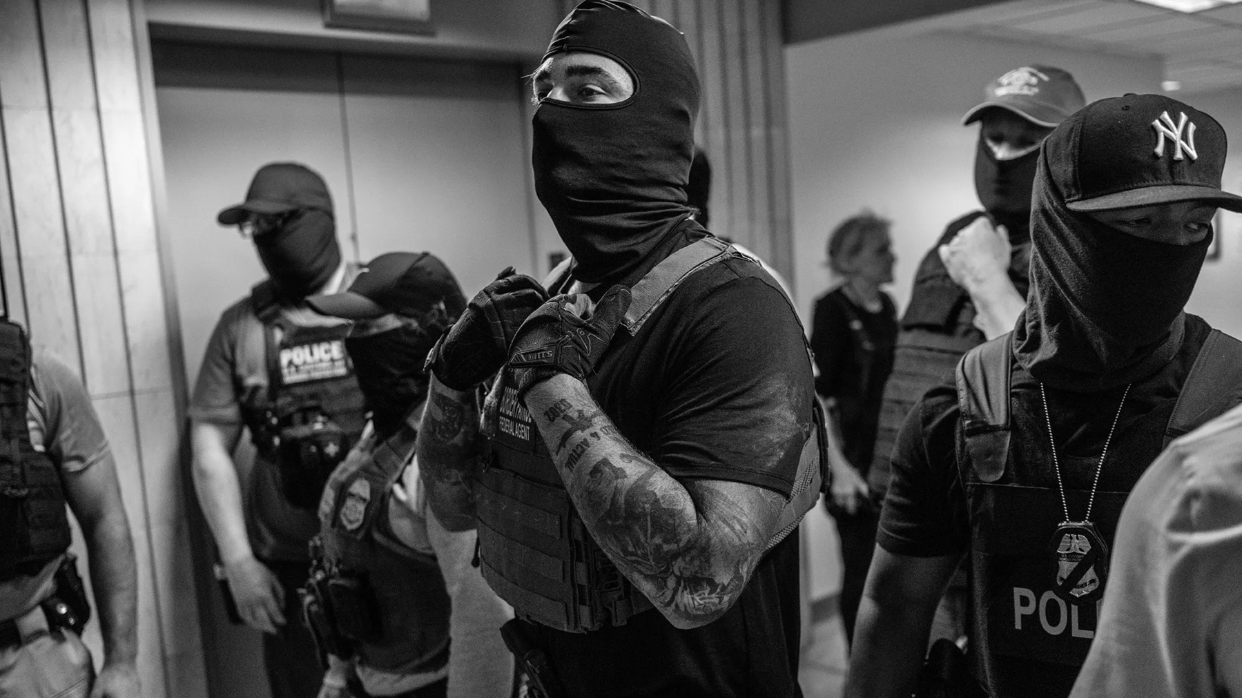 A group of masked individuals, some wearing police vests, stand in a hallway.
