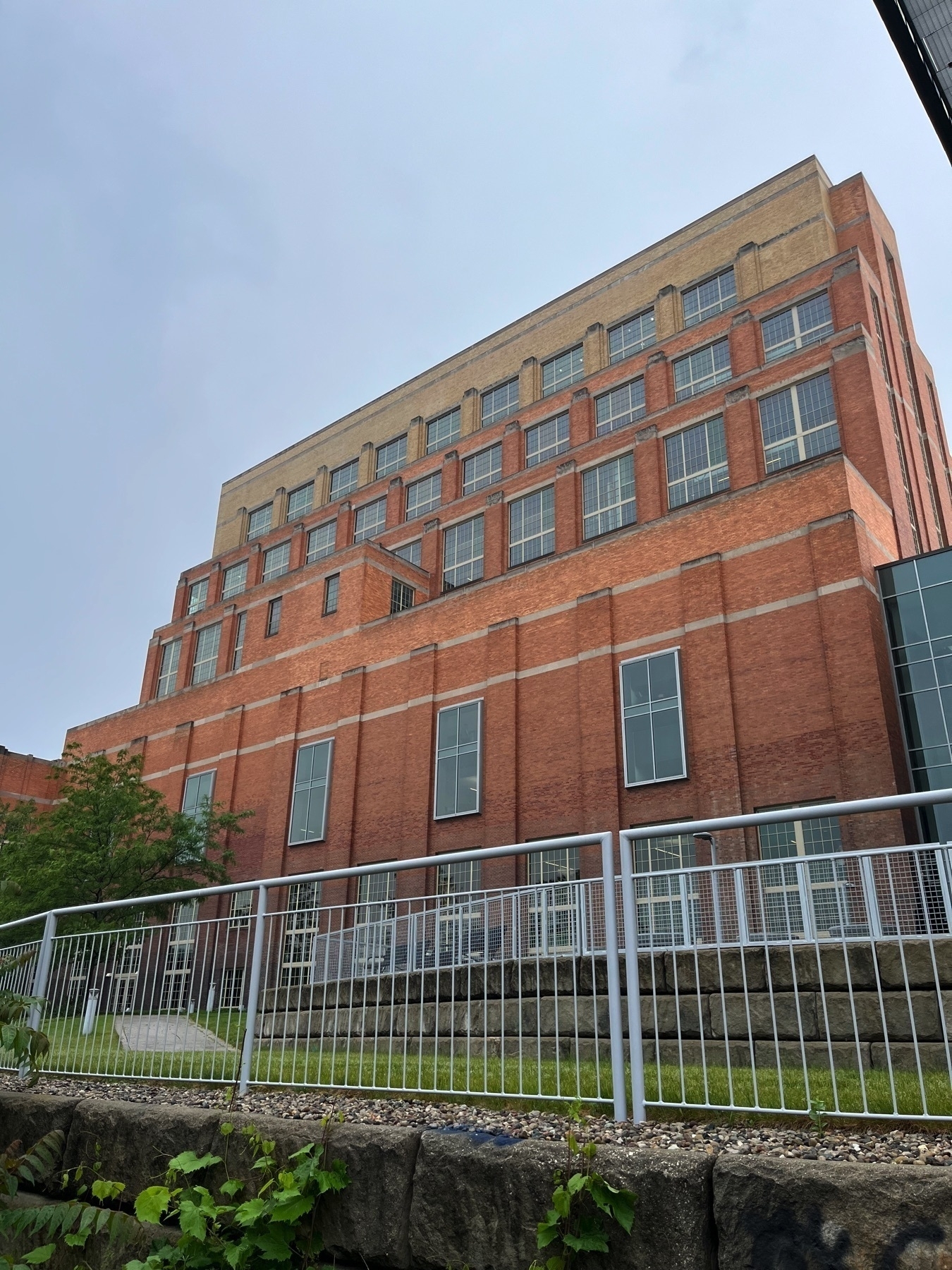A picture of the Ottowa Street Power Station in downtown Lansing. A large brick building with multiple stories and rows of windows is viewed from a low angle, partially obscured by a metal fence and elevated grassy area.
