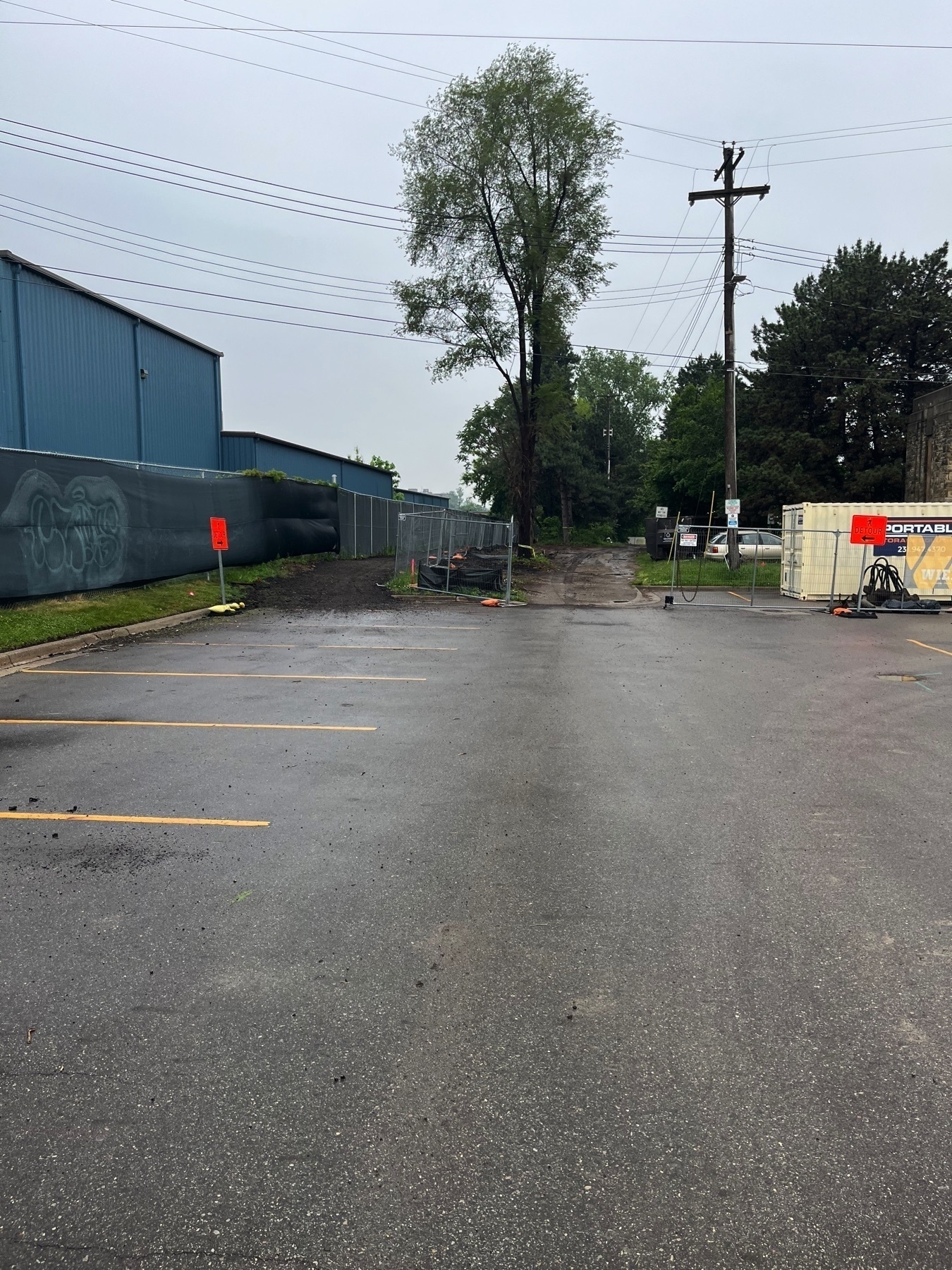 A wet, deserted parking lot is lined with a blue industrial building and a path leading through trees, featuring construction signs and equipment.
