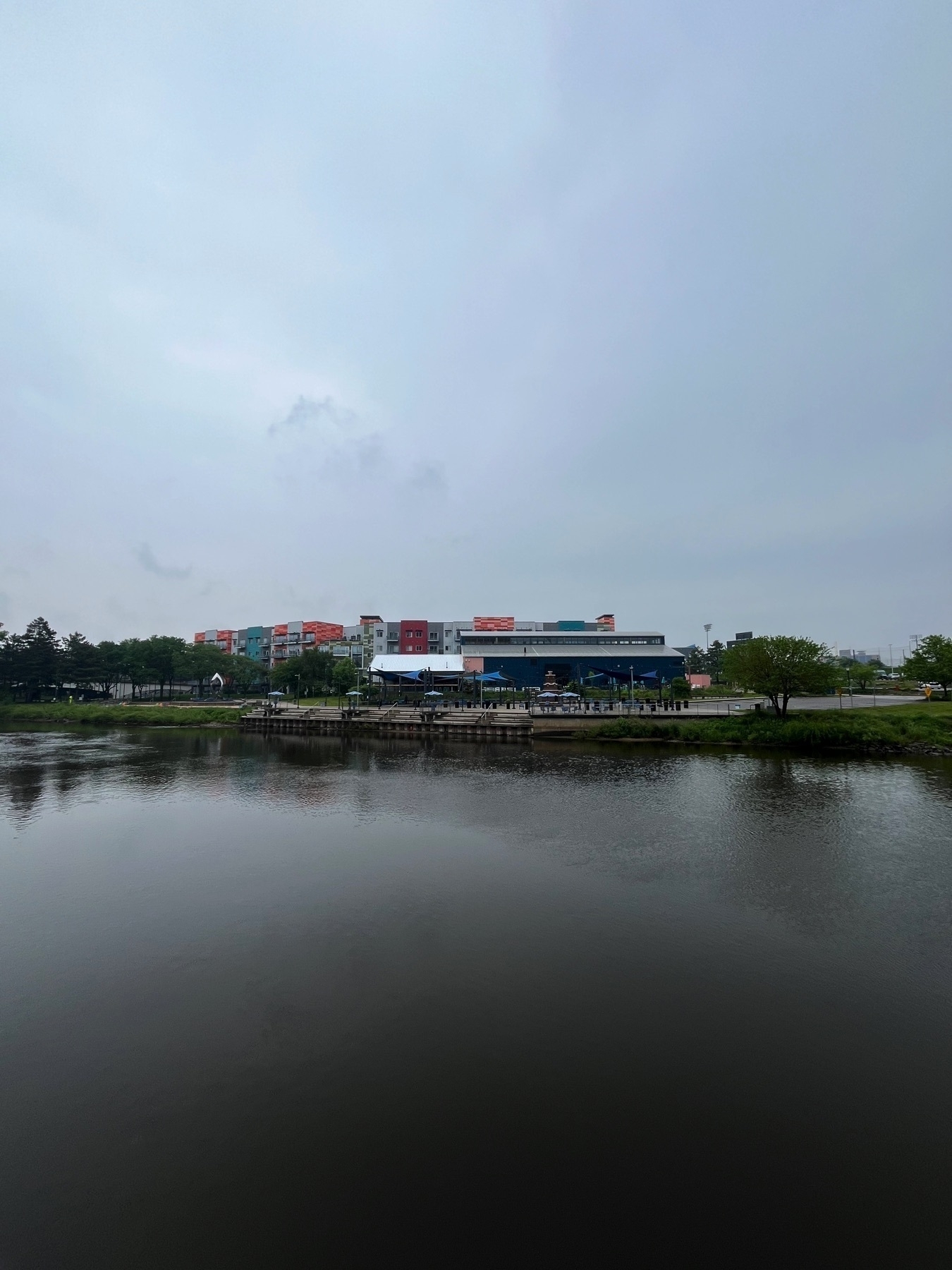 A calm river reflects the overcast sky, with a backdrop of buildings and trees along the riverbank.