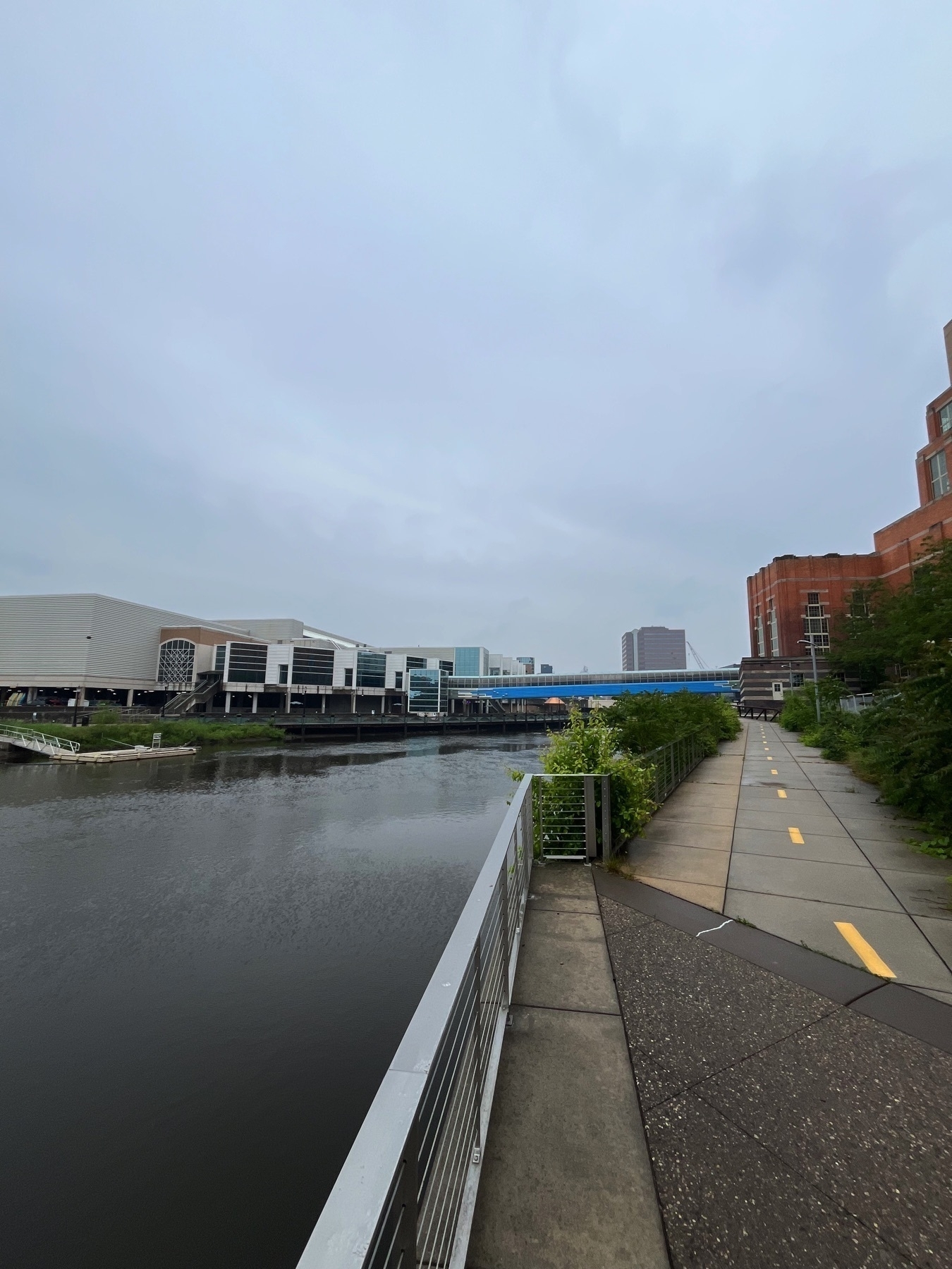 Looking south along the west bank of the Grand River in Lansing. A riverside walkway with a bridge and modern buildings in the background under an overcast sky.