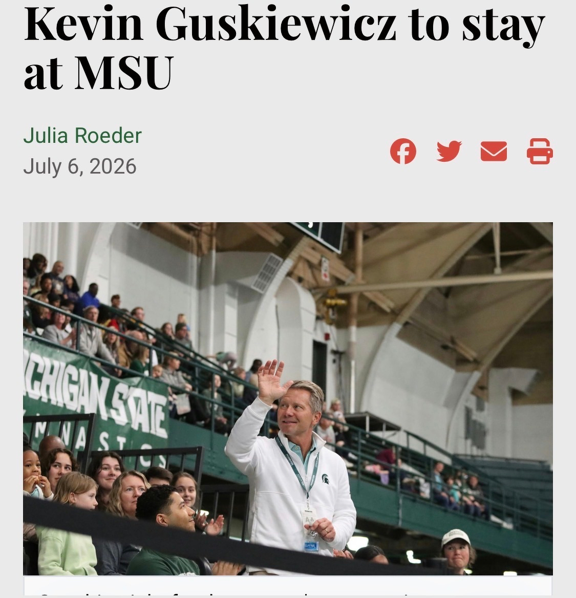A screenshot of a news article from July 6, 2026, by Julia Roeder, titled &ldquo;Kevin Guskiewicz to stay at MSU.&rdquo; The accompanying photograph shows Michigan State University President Kevin Guskiewicz smiling and waving to a crowd inside an indoor athletic arena. He is wearing a white quarter-zip pullover featuring a green Spartan logo on the chest and an event credential lanyard around his neck. In the background, spectators look on from green stadium seating behind a railing featuring a &ldquo;MICHIGAN STATE GYMNASTICS&rdquo; sign.