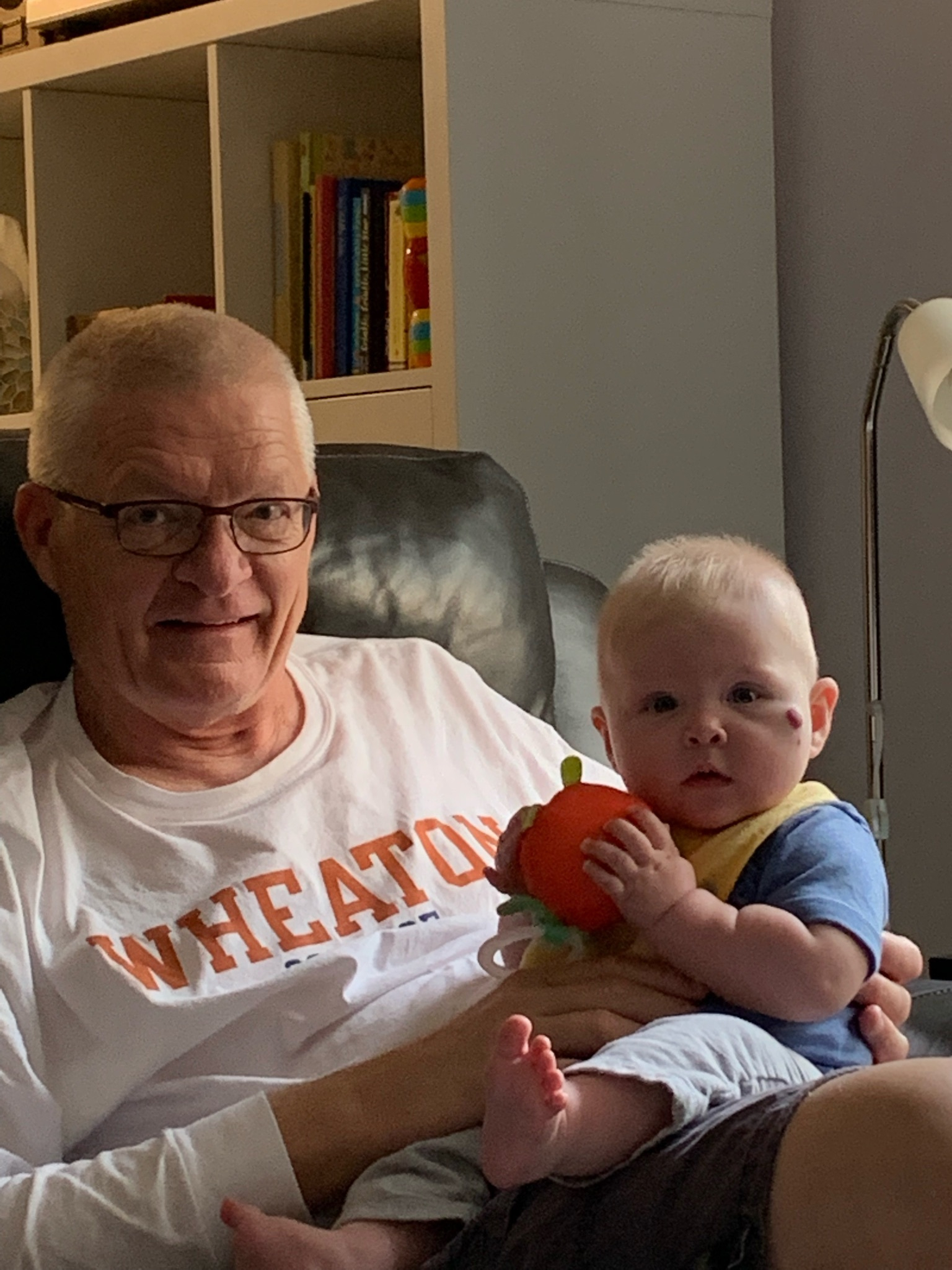A man wearing glasses and a Wheaton shirt is sitting on a couch holding a baby who is clutching a small orange toy.