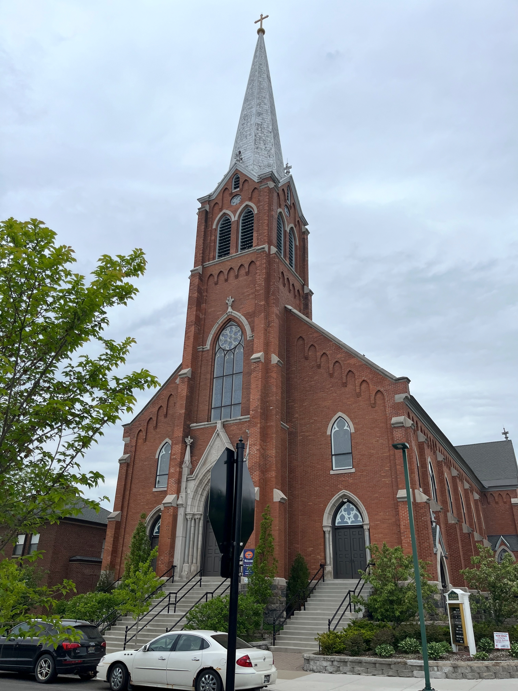 A red brick church with a tall steeple against a grey cloudy sky framed by a tree on its left side.