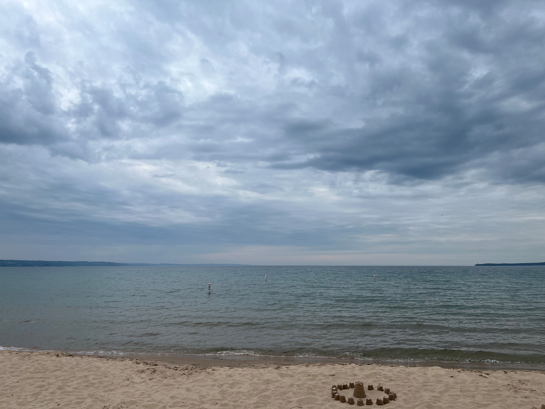 Cloudy skies stretch over a calm beach with gentle waves and a small sand structure in the foreground.