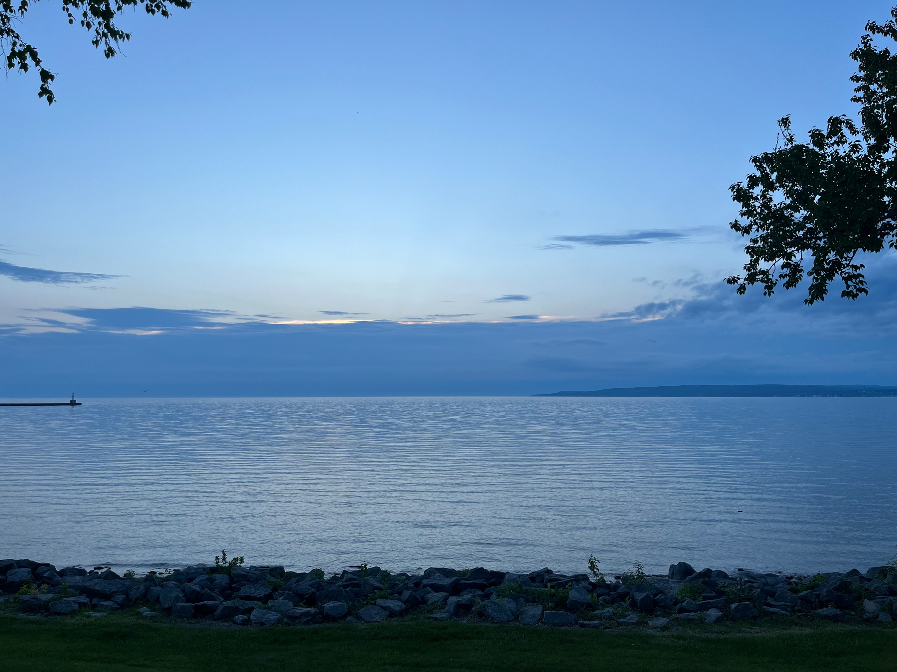A tranquil lakeside scene at dusk features calm waters, a rocky shoreline, and silhouettes of trees under a pale blue sky.