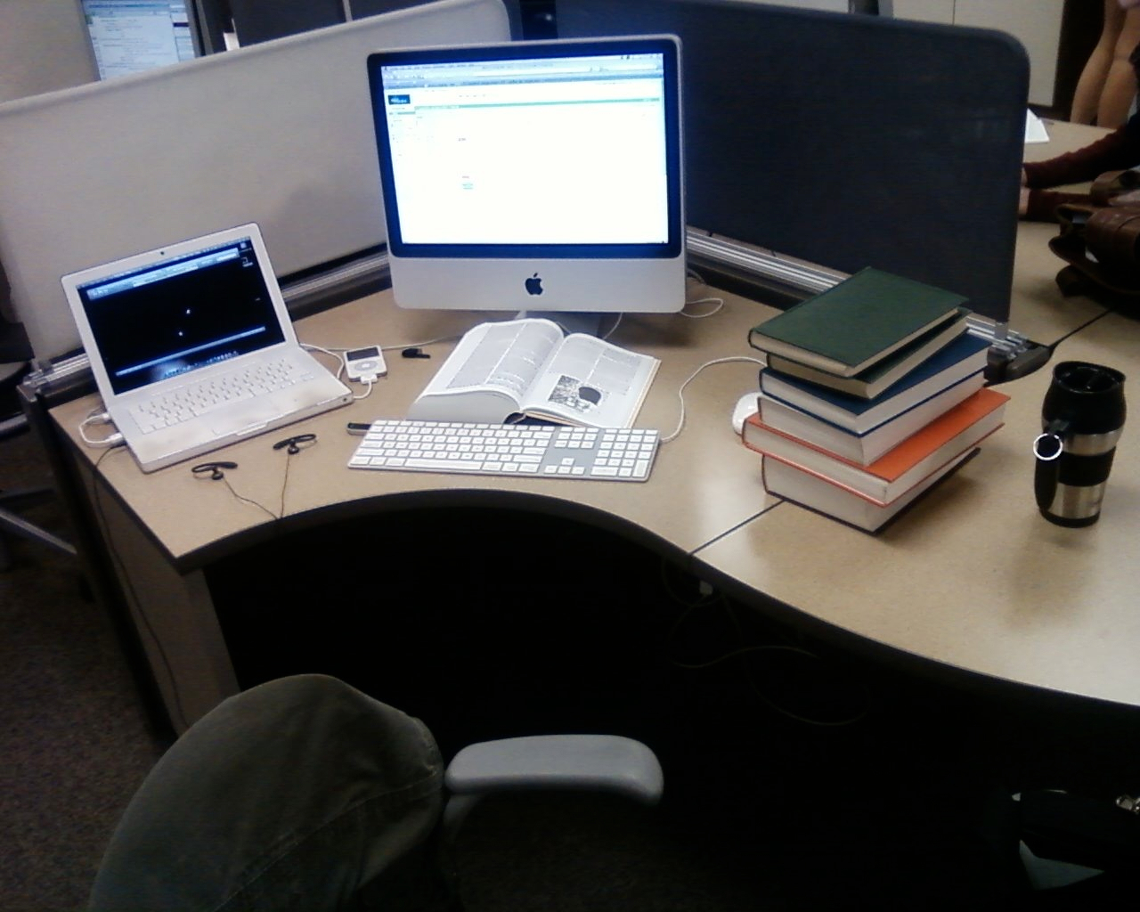 Auto-generated description: A workspace featuring a desktop computer, a laptop, several open and closed books, a tumbler, and scattered items.
