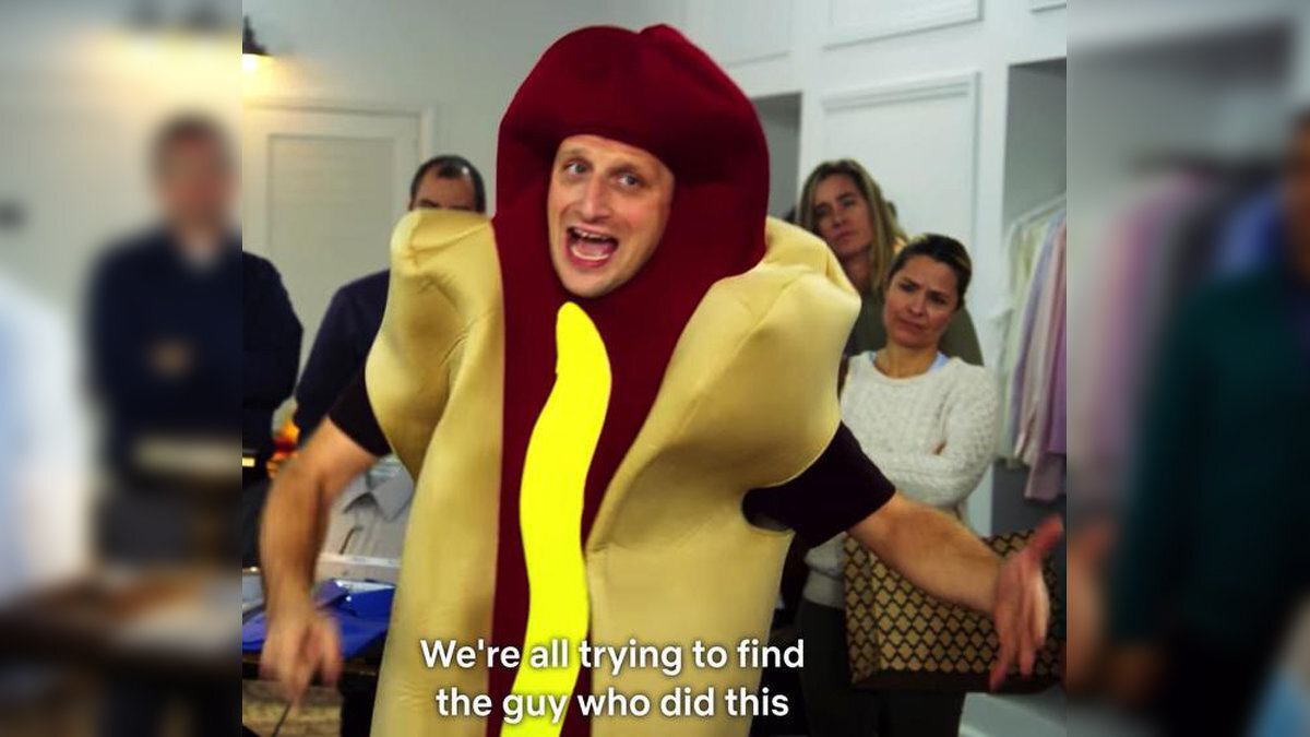 A person dressed in a hot dog costume is speaking animatedly in a room with several people watching, accompanied by the caption We&rsquo;re all trying to find the guy who did this.