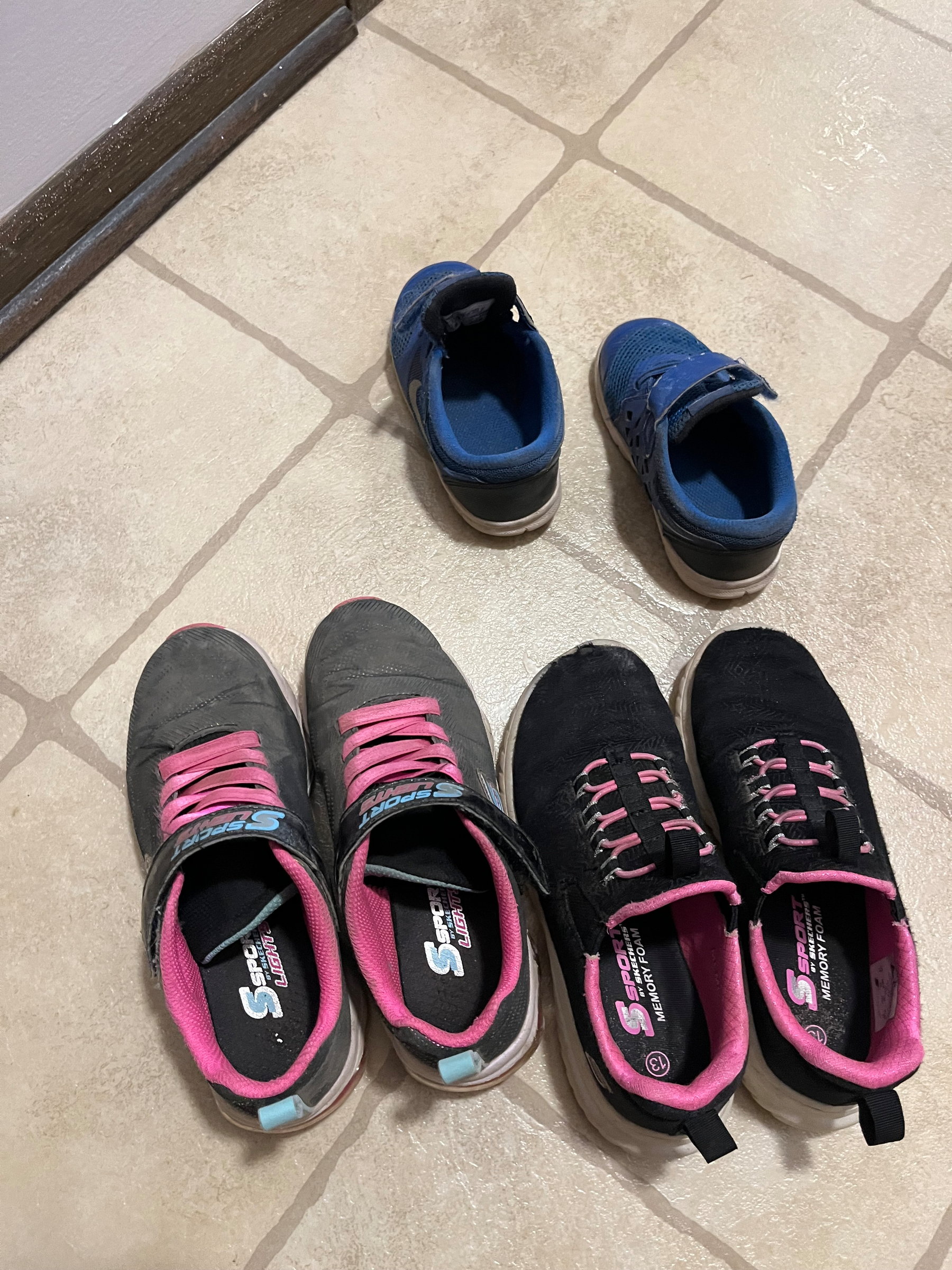 Three pairs of shoes, two with black and pink accents and one with blue and black, are placed on a tiled floor near a wall.