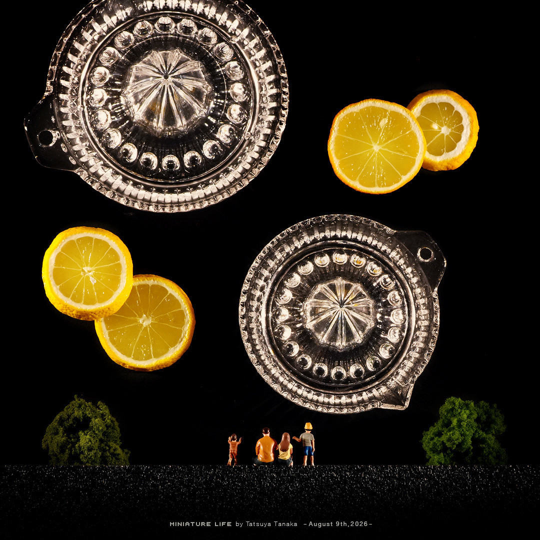 A miniature diorama by Tatsuya Tanaka for August 9th, 2026. Against a dark black background, two glass citrus squeezers and bright yellow lemon slices are arranged in the sky to resemble sparkling, illuminated fireworks. At the bottom, a tiny family figurine set—two parents and two children—sits and stands between two miniature trees, gazing up at the glowing lemon and glass display.
