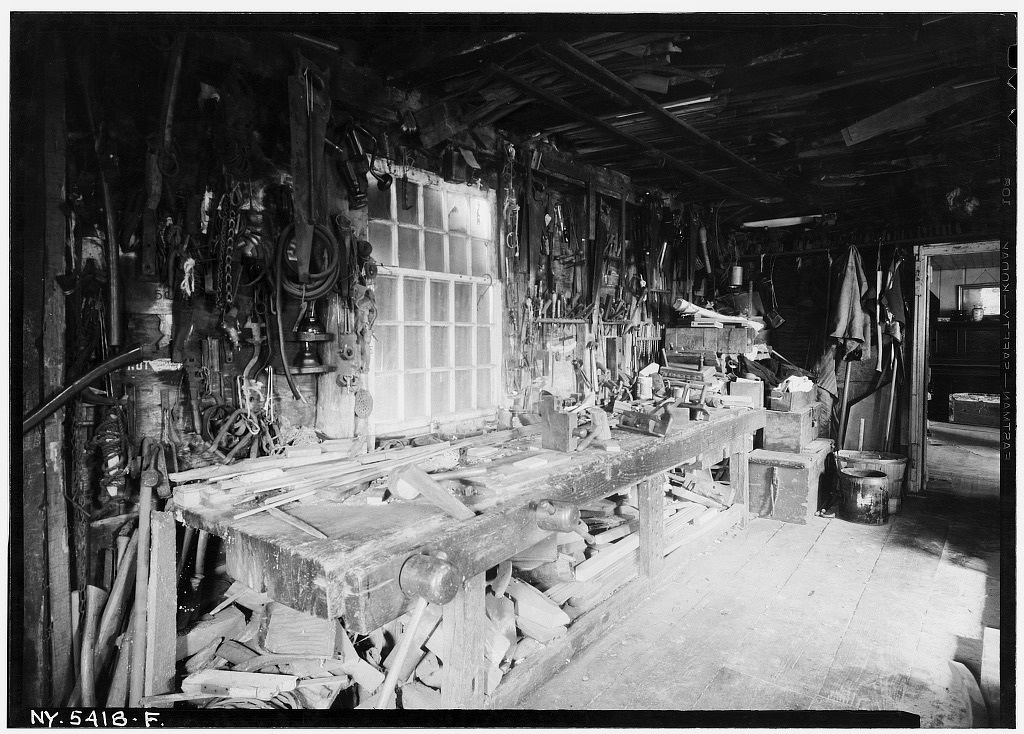 interior of carpenter shop. View from north end. - Dominy House, North Main Street, East Hampton, Suffolk County, NY. Photo by Stanley P. Mixon, July 20, 1940. [hdl.loc.gov/loc.pnp/h...](http://hdl.loc.gov/loc.pnp/hhh.ny0772/photos.123503p.) A historic black-and-white interior photograph (labeled NY-5418-F) depicting a densely packed traditional carpenter's workshop. A long, heavy wooden workbench sits beneath a large multi-pane window, covered in hand planes, chisels, wooden blocks, and hand tools. Dozens of hand saws, clamps, and various woodworking tools hang densely on the exposed timber walls.
