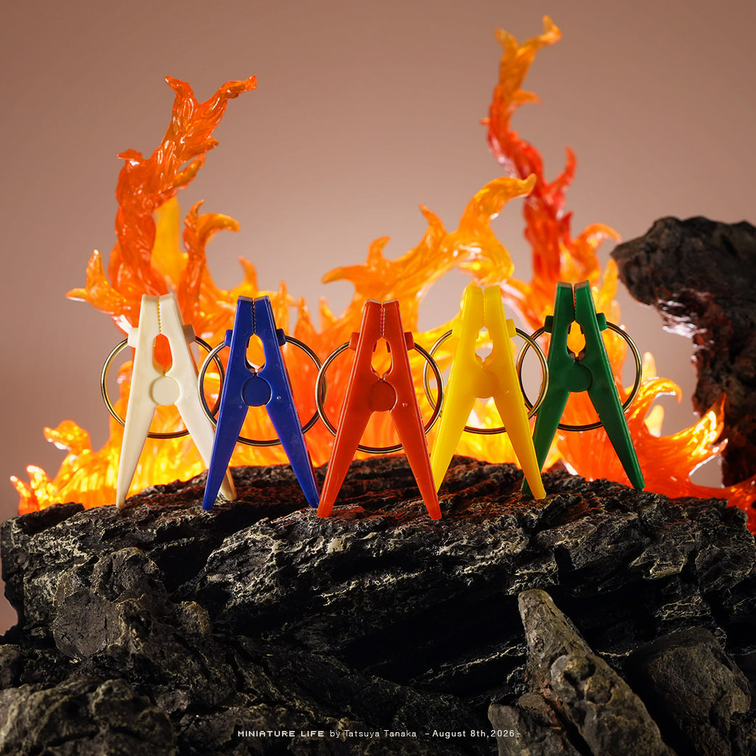 A superhero-themed miniature diorama by Tatsuya Tanaka for August 8th, 2026. Five colorful plastic clothespins—white, blue, red, yellow, and green—are lined up atop rugged black volcanic rock, posed like a classic Sentai or Power Rangers hero team. Behind them, vibrant, translucent orange and red flame effects create a dramatic, fiery explosion backdrop.