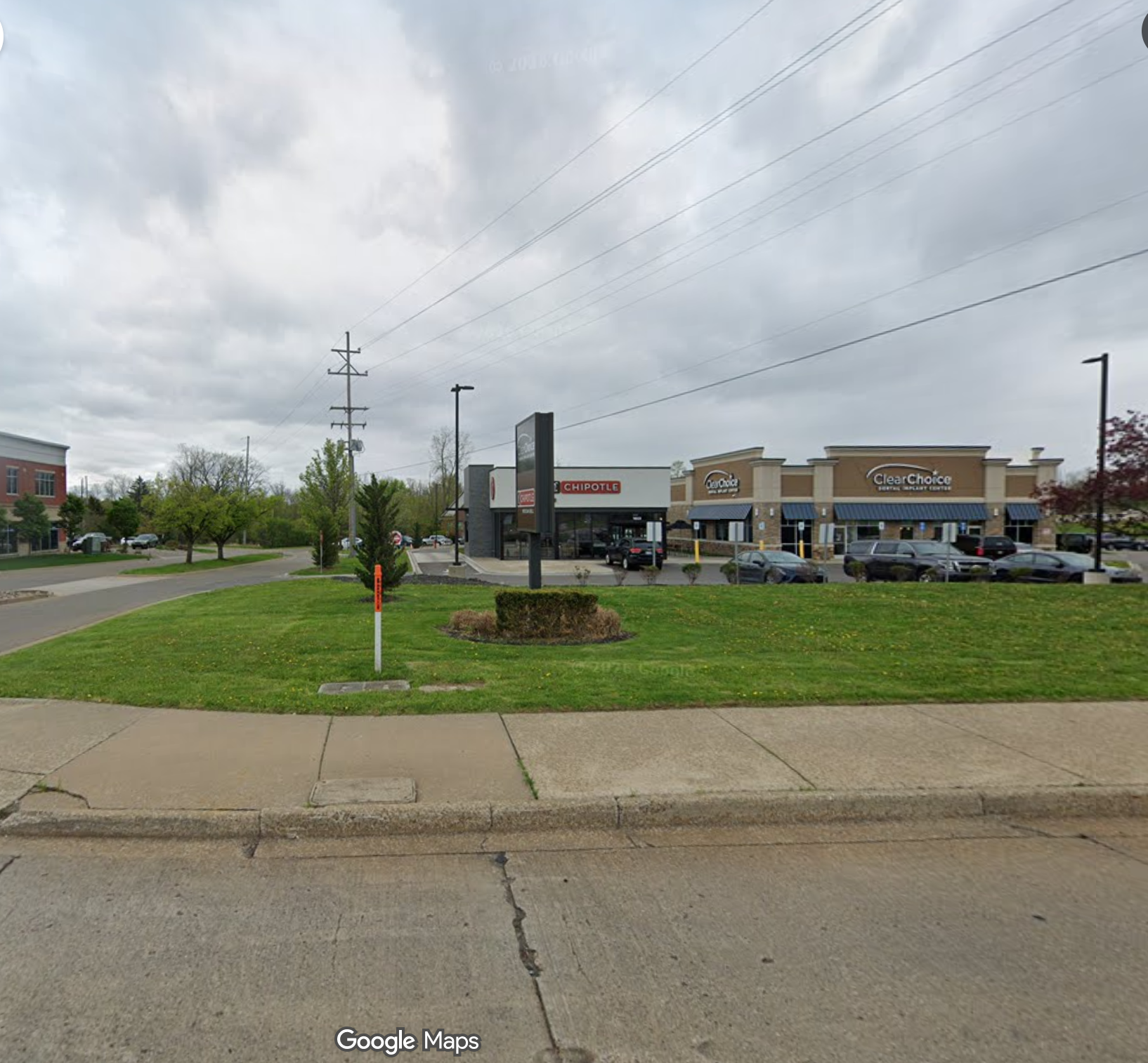 A Google Maps street view screenshot of a commercial plaza under an overcast sky. In the background are standalone retail storefronts for a Chipotle Mexican Grill and a ClearChoice Dental Implant Center. In the foreground, a manicured green lawn with a small landscaped island sits along a concrete sidewalk and asphalt street.